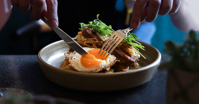 A person cutting into a plated dish featuring a fried egg, greens, and corned beef