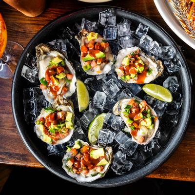 Raw oysters on ice topped with avocado, tomato, and sauce, garnished with lime wedges.