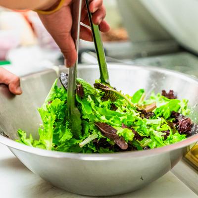 A hand with tongs tosses mixed greens in a large bowl.