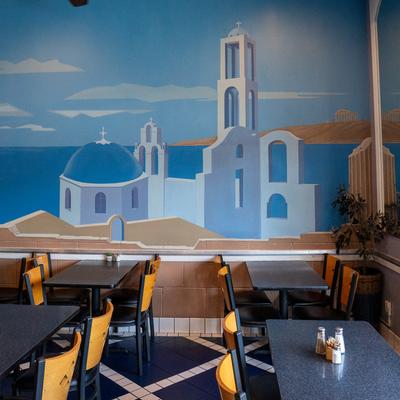 Diner tables, chairs, painted wall behind with greek sea view and white otrodox church.