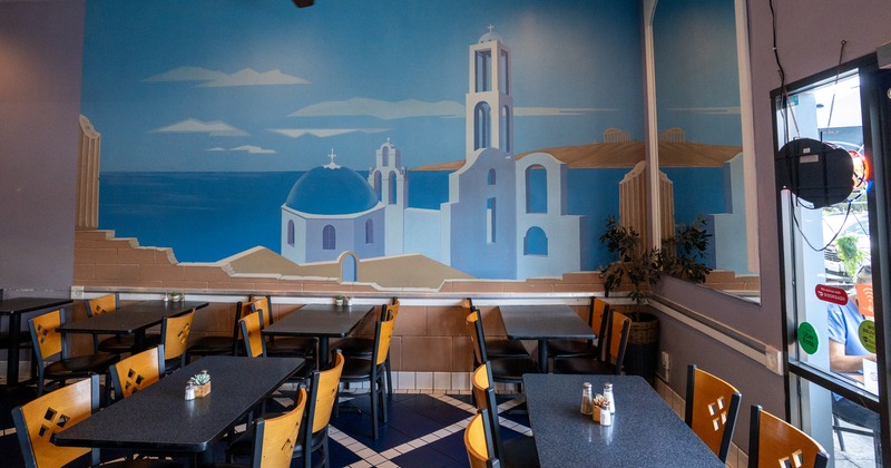 Diner tables, chairs, painted wall behind with greek sea view and white otrodox church