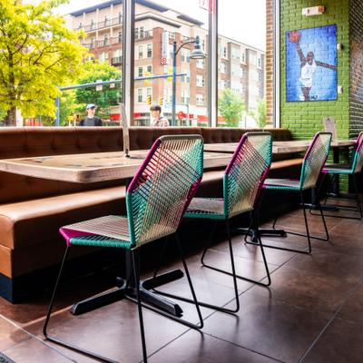 Booth seating with tables and multicolored woven chairs next to a window.