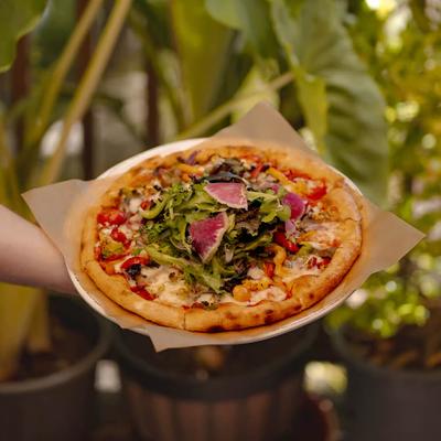 Hand holding a wood-fired pizza topped with a fresh salad.