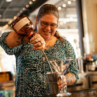 A smiling bartender pours liquor into a shaker at a bar.