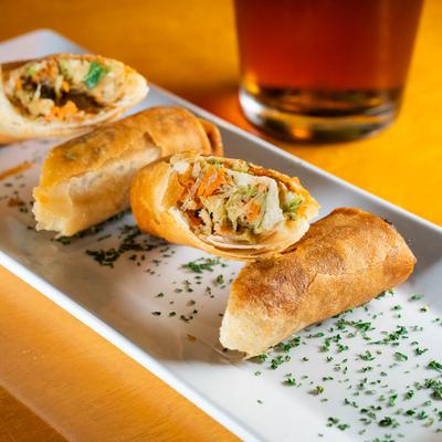 A white rectangular plate holds Southwest Eggrolls, a glass of dark beer sits nearby.