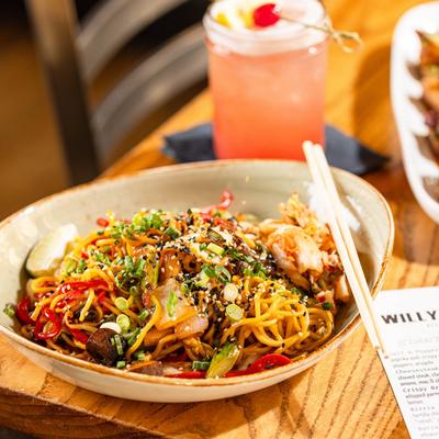 Stir-fried noodles topped with vegetables, sesame seeds, and scallions, served with a cocktail.