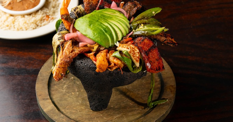 Molcajete filled with various meats and seafood, topped with sliced avocado