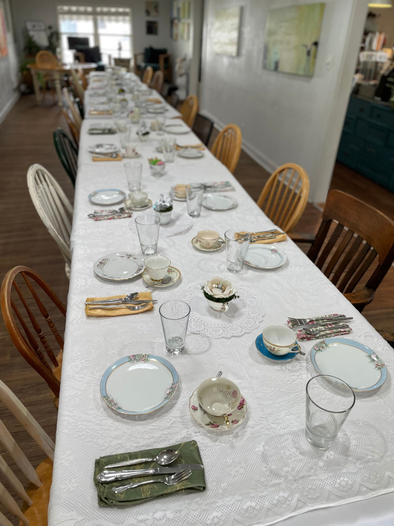 A set table with a lace tablecloth, vintage tea cups, plates, and silverware, awaiting guests.