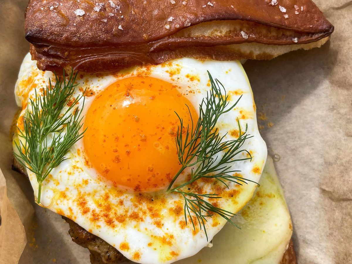 A sunny-side fried egg with a sprig of a green herb and dots of sauce on top of a croissant slice.