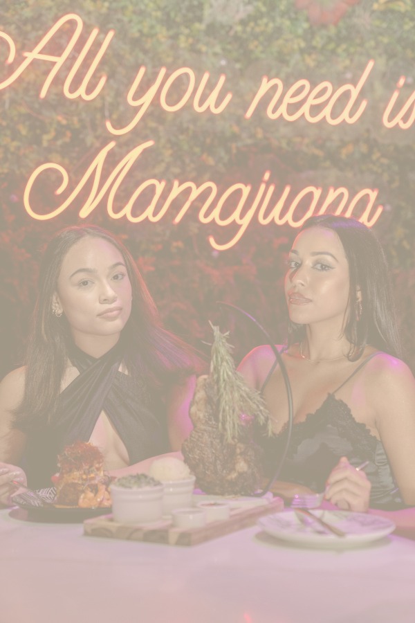 Two women sitting at a table with a neon sign that says all you need is marijuana.