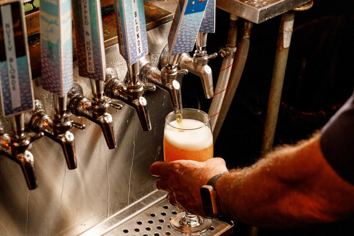 Hand pouring pale beer from a metal tap into a foamy pint glass at a bar