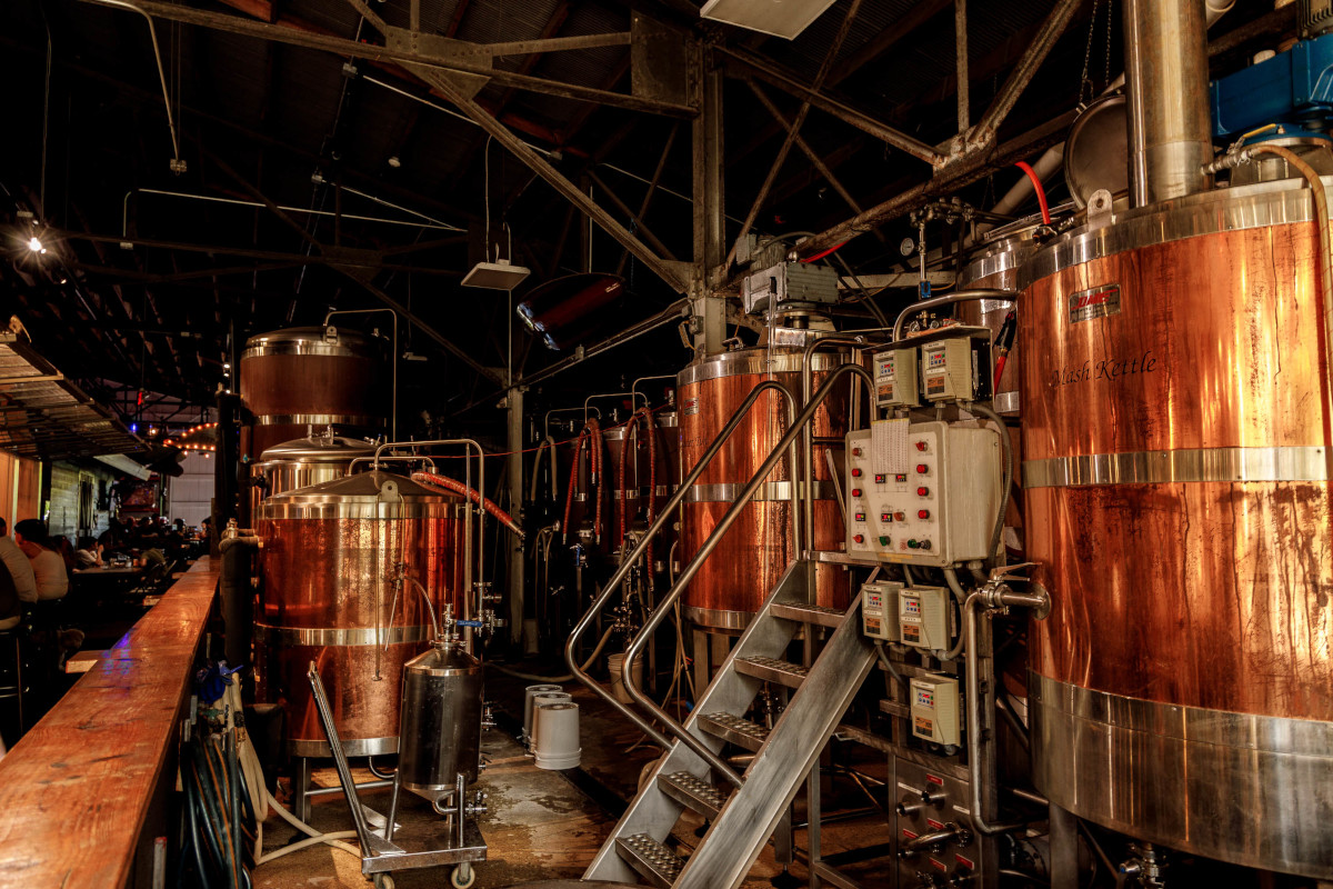 Interior of a brewery with large copper brewing tanks, metal pipes, control panels, and a staircase