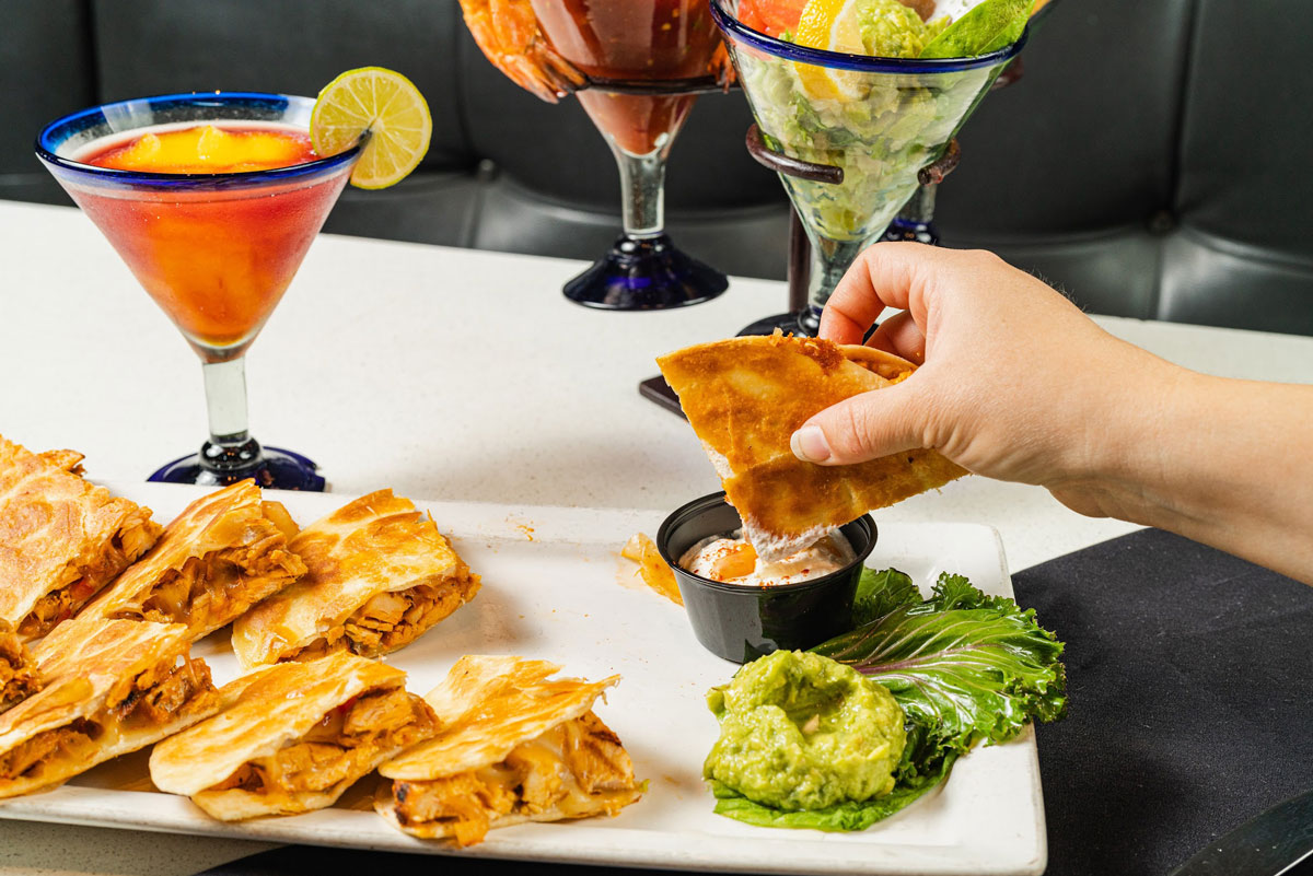 Hand dipping chicken quesadilla in a sauce, cocktails in the background