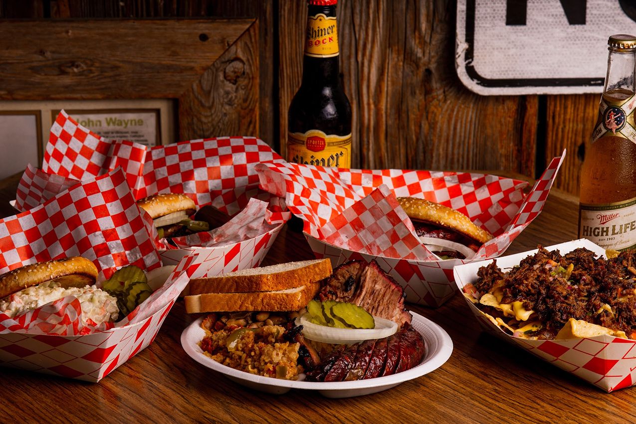 BBQ catering trays with brisket, ribs, sandwiches, and sides at Augie’s Barbed Wire Smokehouse in San Antonio, displayed with Shiner Bock and Miller High Life.