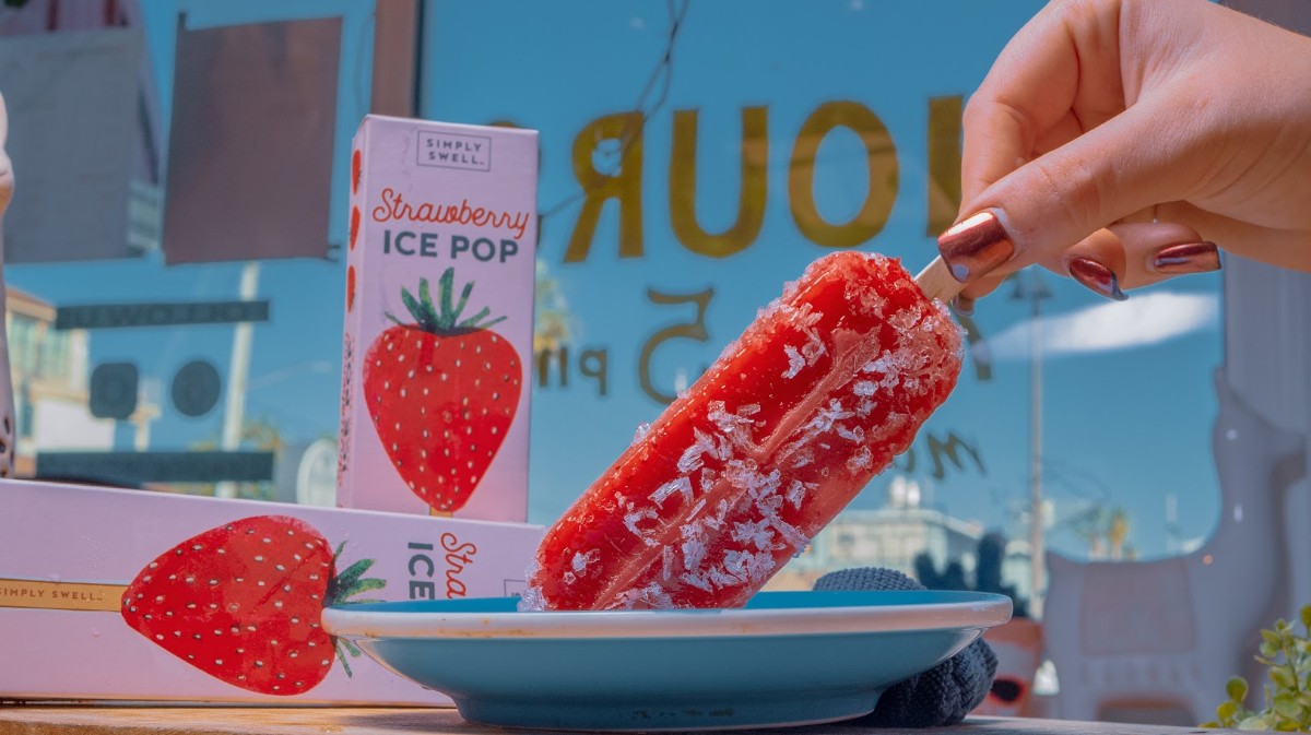 Side view of hand holding a strawberry ice pop with ice pop boxes in background