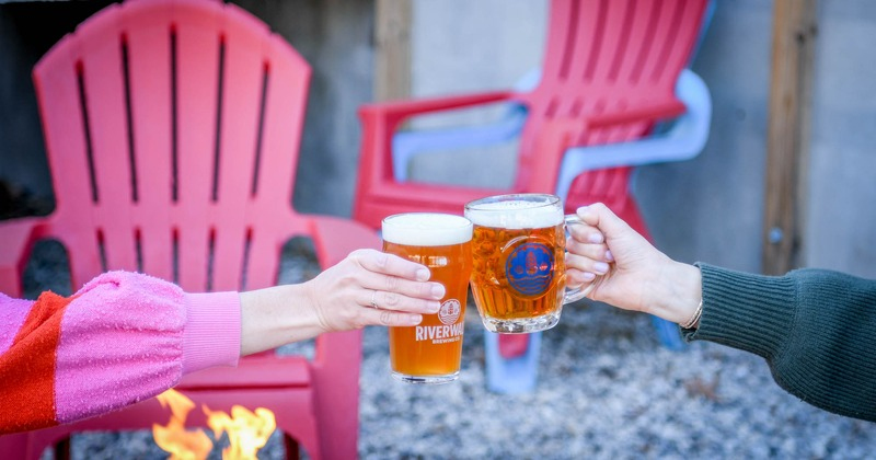 Two people toasting beer glasses in front of a fire pit.