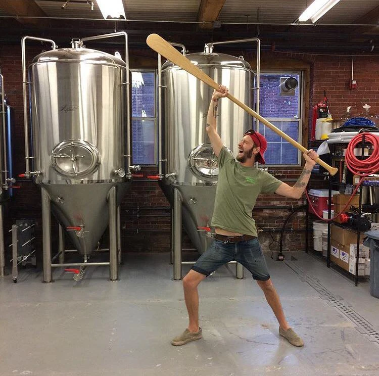 Stevie raising a wooden mixing paddle at the brewery tanks