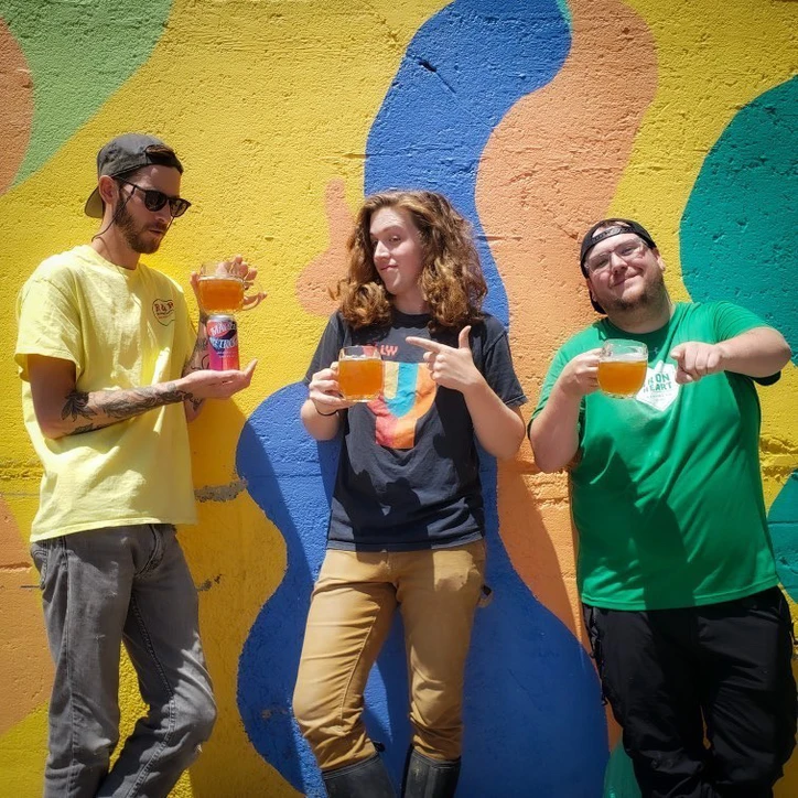 Team members posing with beers in front of a colorful mural