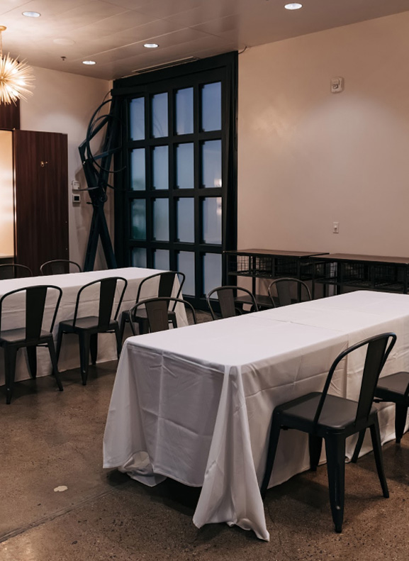 Empty dining area with tables covered in white tablecloths and black chairs near a large window.