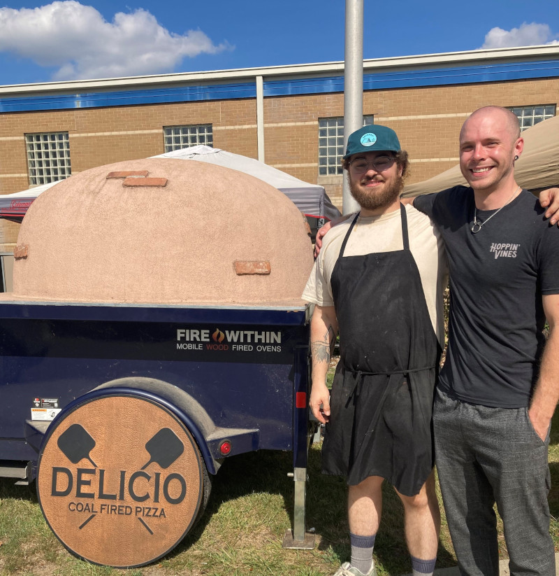 two men standing next to a pizza trailer
