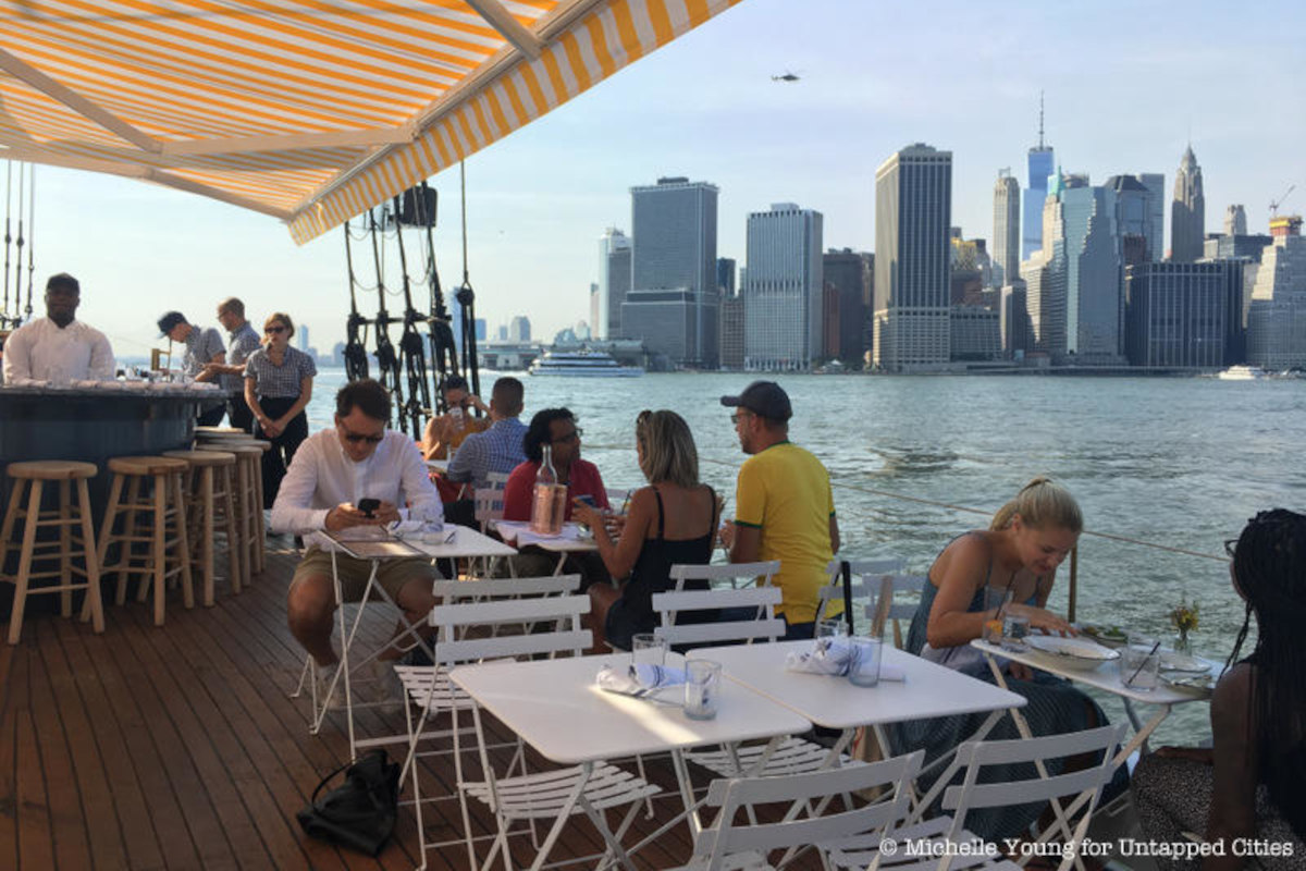 a group of people sitting at tables on a boat with a view of manhattan.