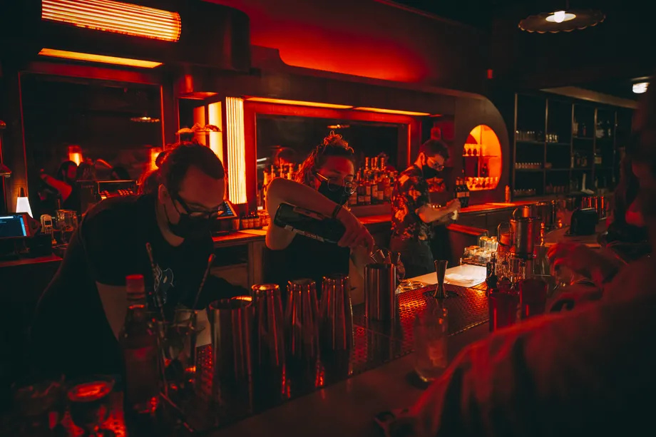 Bartenders wearing masks pour drinks behind a bar lit with red lights.