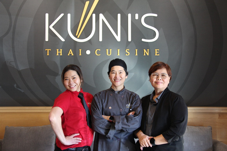 Three people are posing in front of Kunis Thai Cuisine restaurant sign.