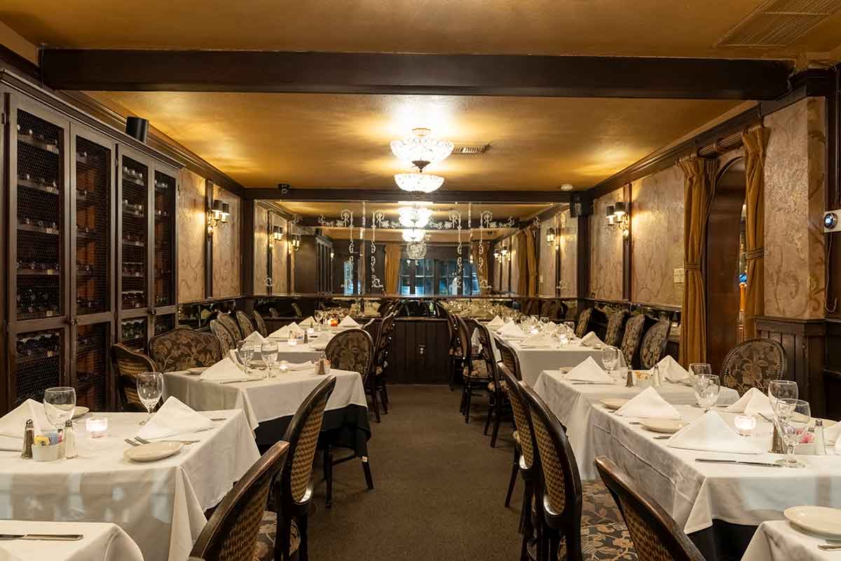 A classic restaurant dining room with set tables, patterned chairs, and a large mirror.