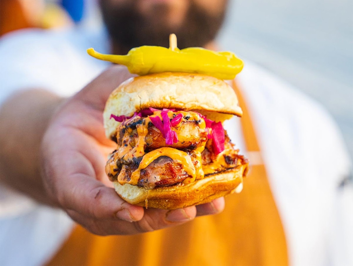A close-up of a sandwich with grilled filling, purple cabbage, and a pickled pepper on top