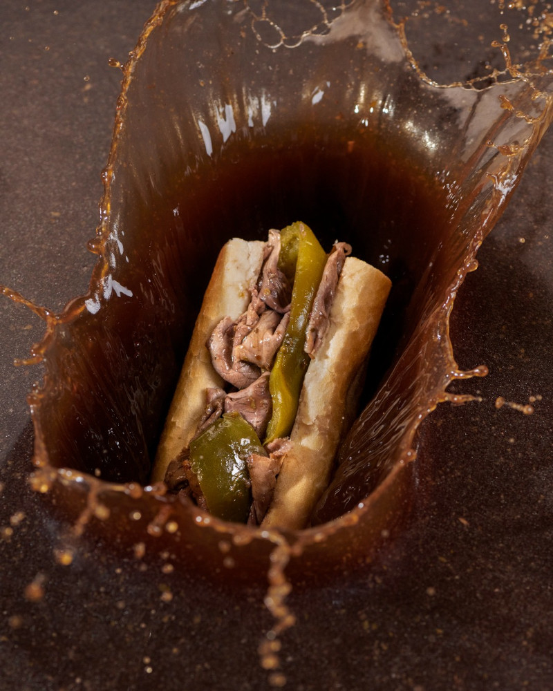 A sandwich with meat and green peppers is surrounded by a splash of brown liquid.