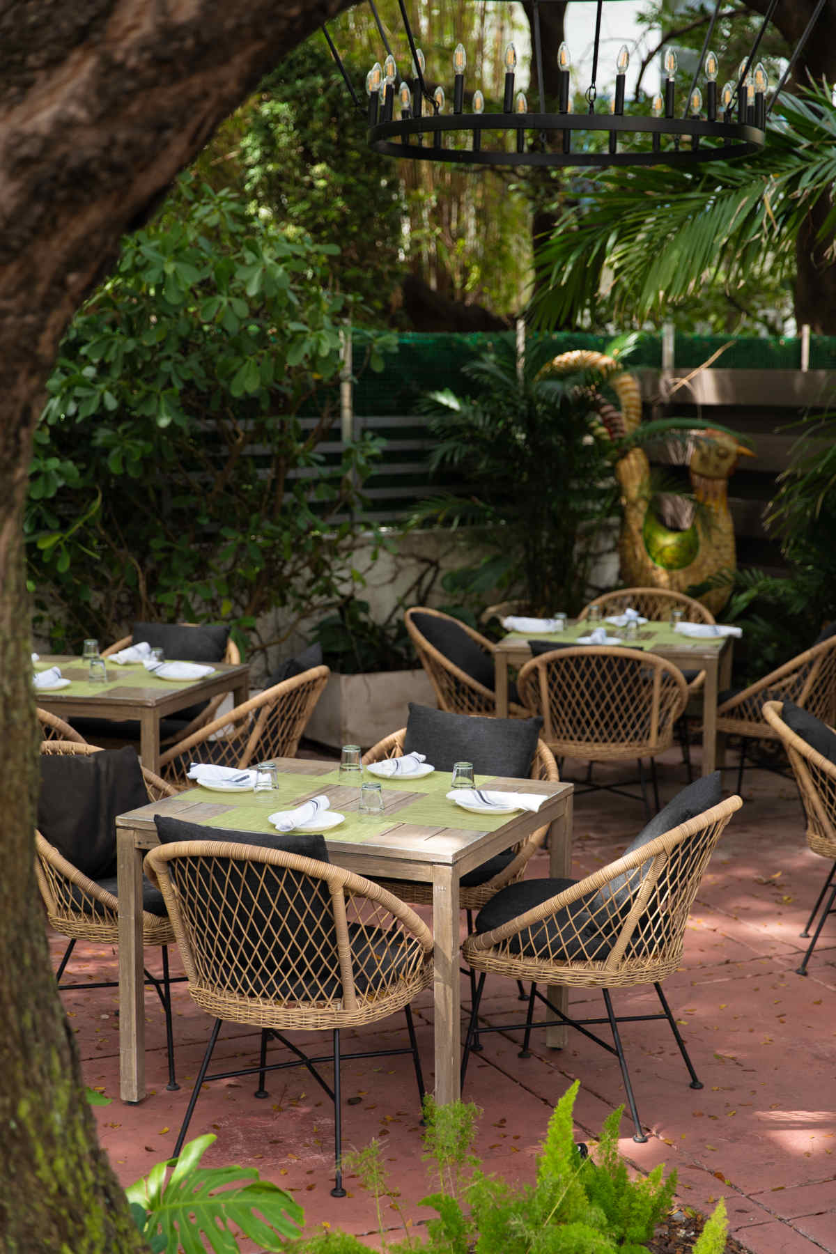 Outdoor restaurant with wooden tables and wicker chairs, surrounded by greenery and trees.