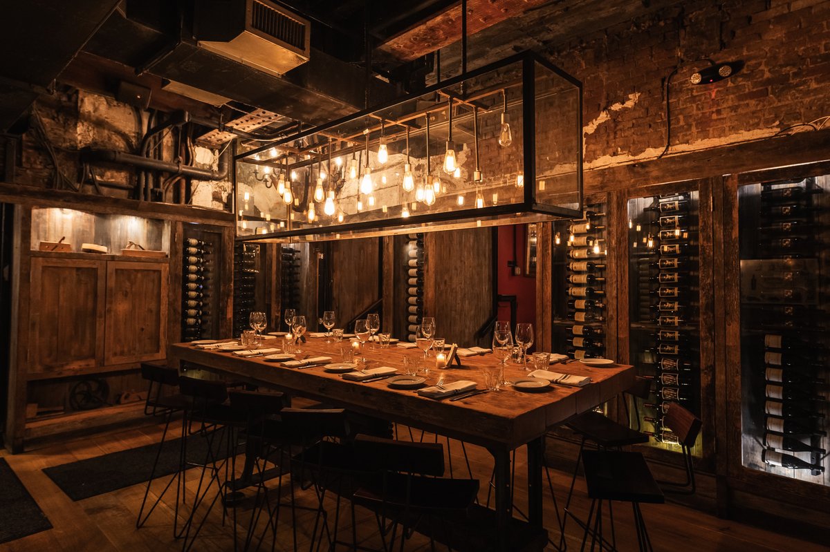 A cozy, dimly lit restaurant with a wooden table set for dinner, pendant lights, and a wine rack.