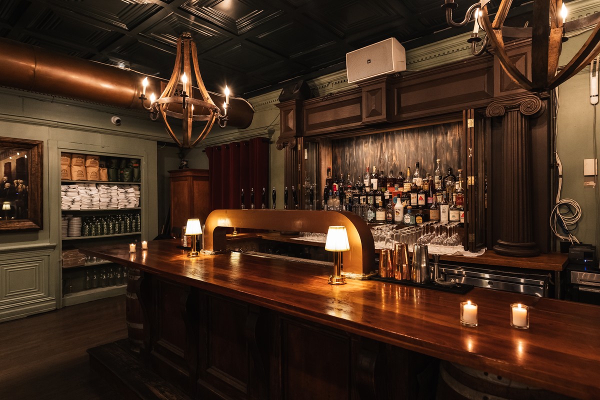 Bar with a wooden counter, stocked shelves, small lamps, and a large chandelier.
