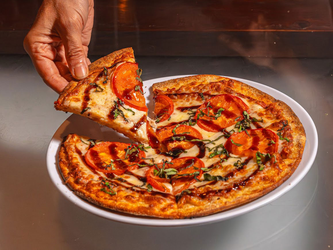 A hand lifting a slice of margherita pizza with fresh tomatoes and basil.
