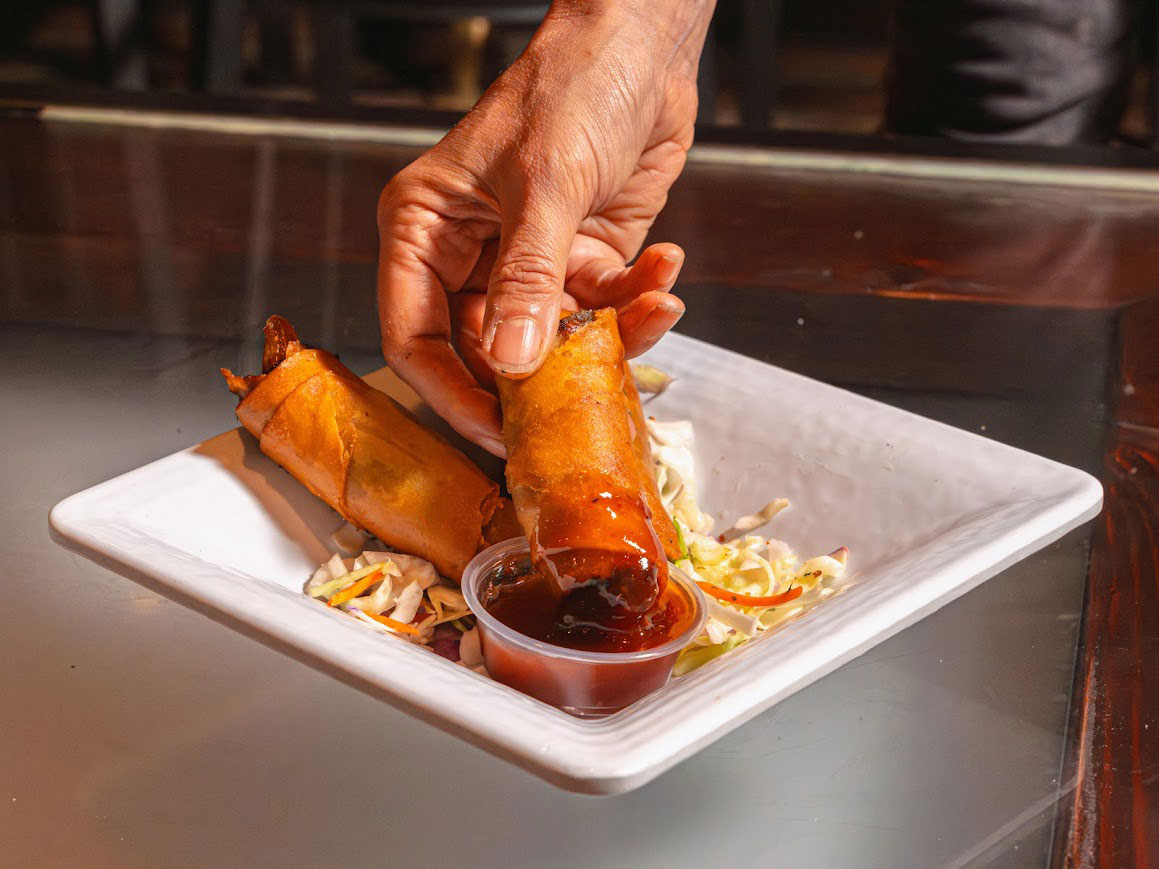 A hand dipping a crispy spring roll into a container of sweet and sour sauce.