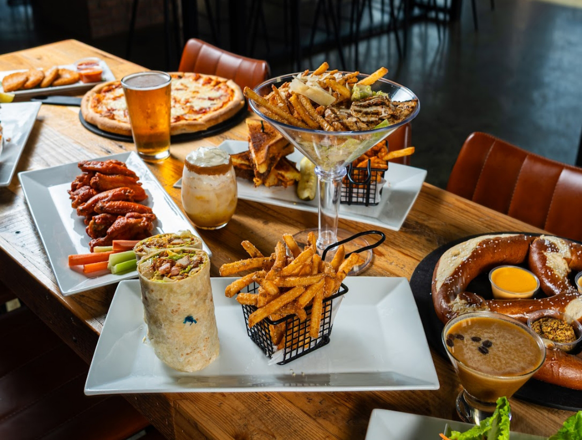 A photo of a food spread from Moonfish Brewery, featuring martini glass of caesar salad, sweet potato fries, buffalo wings, bavarian pretzel with cheese, and a pizza.