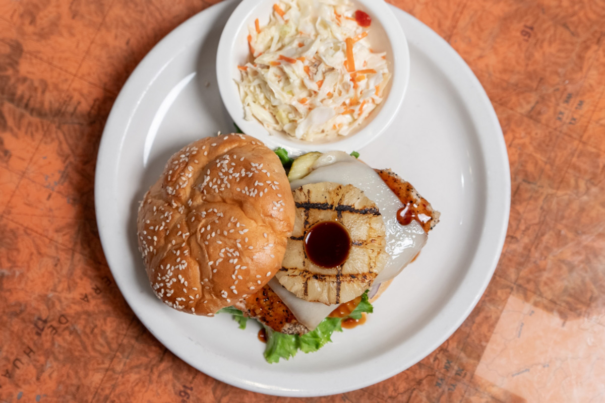 A chicken sandwich with pineapple and cheese on a white plate, accompanied by coleslaw