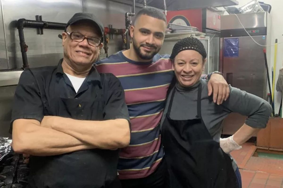 Three people, two in aprons, smile in a commercial kitchen.