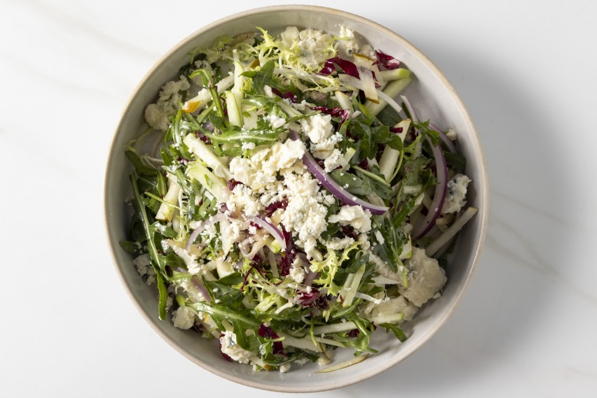 Overhead view of a mixed green salad with white cheese, pale strips, and purple onion in a bowl