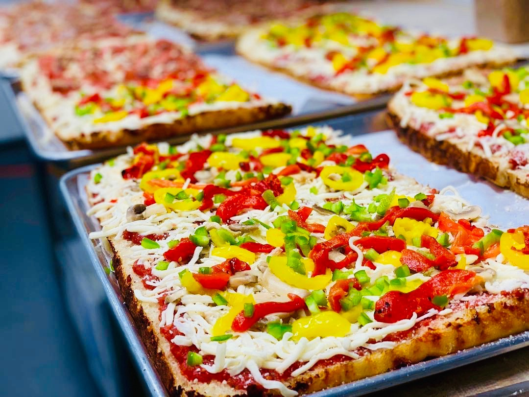 Rectangular pizzas topped with vibrant red, yellow, and green peppers, mushrooms, and melted cheese on trays