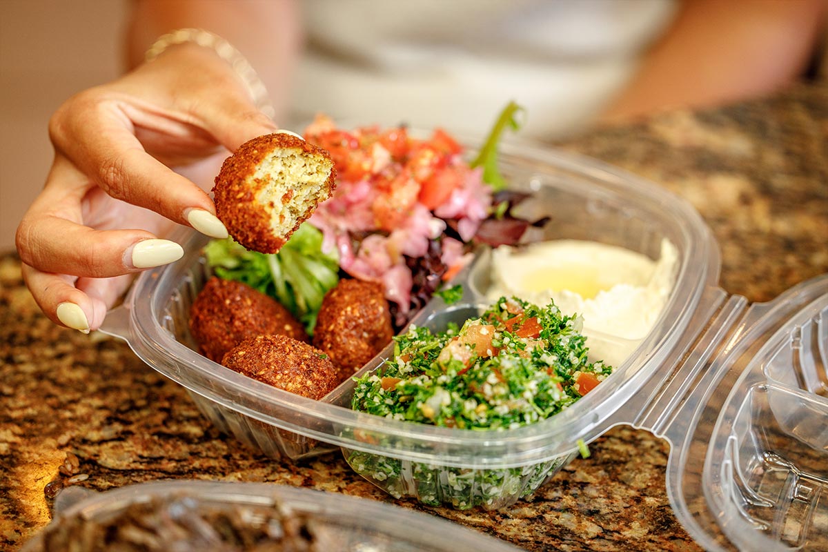 Hand holding falafel over takeout box with tabbouleh and hummus