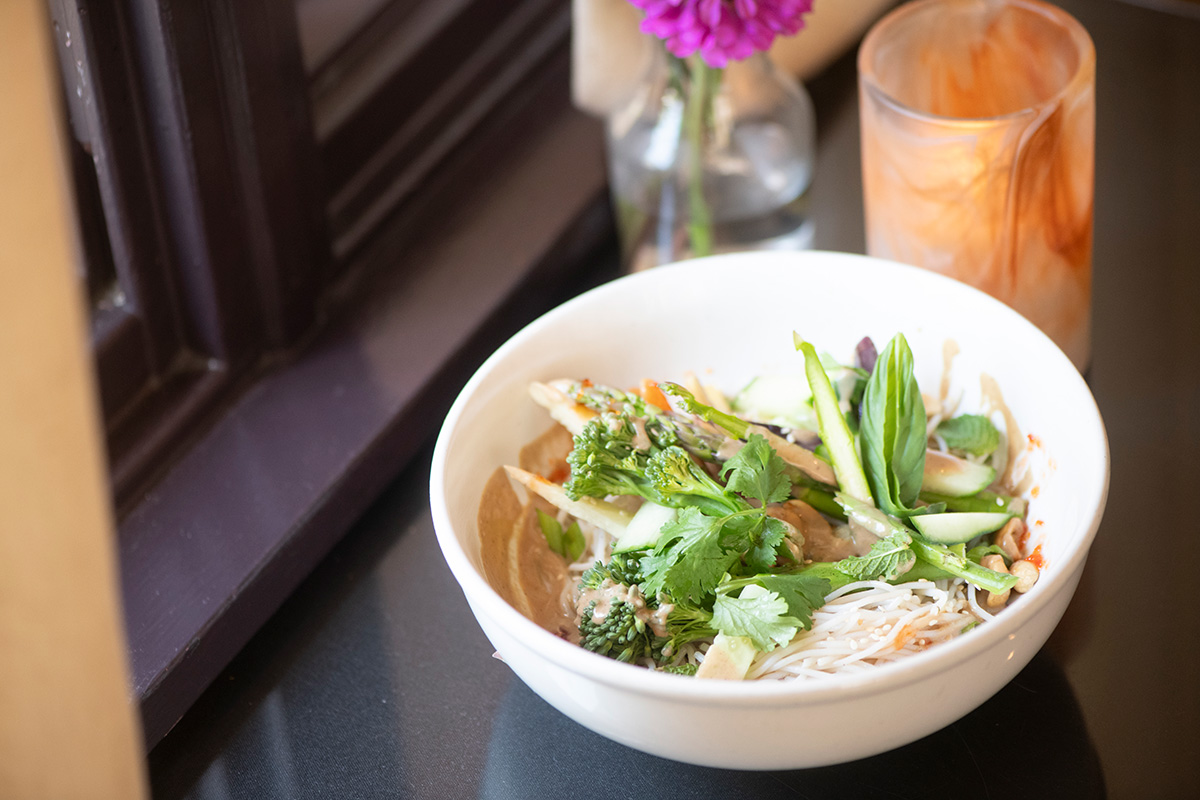 Bowl of noodles and fresh vegetables on a dark table beside a vase of purple flowers