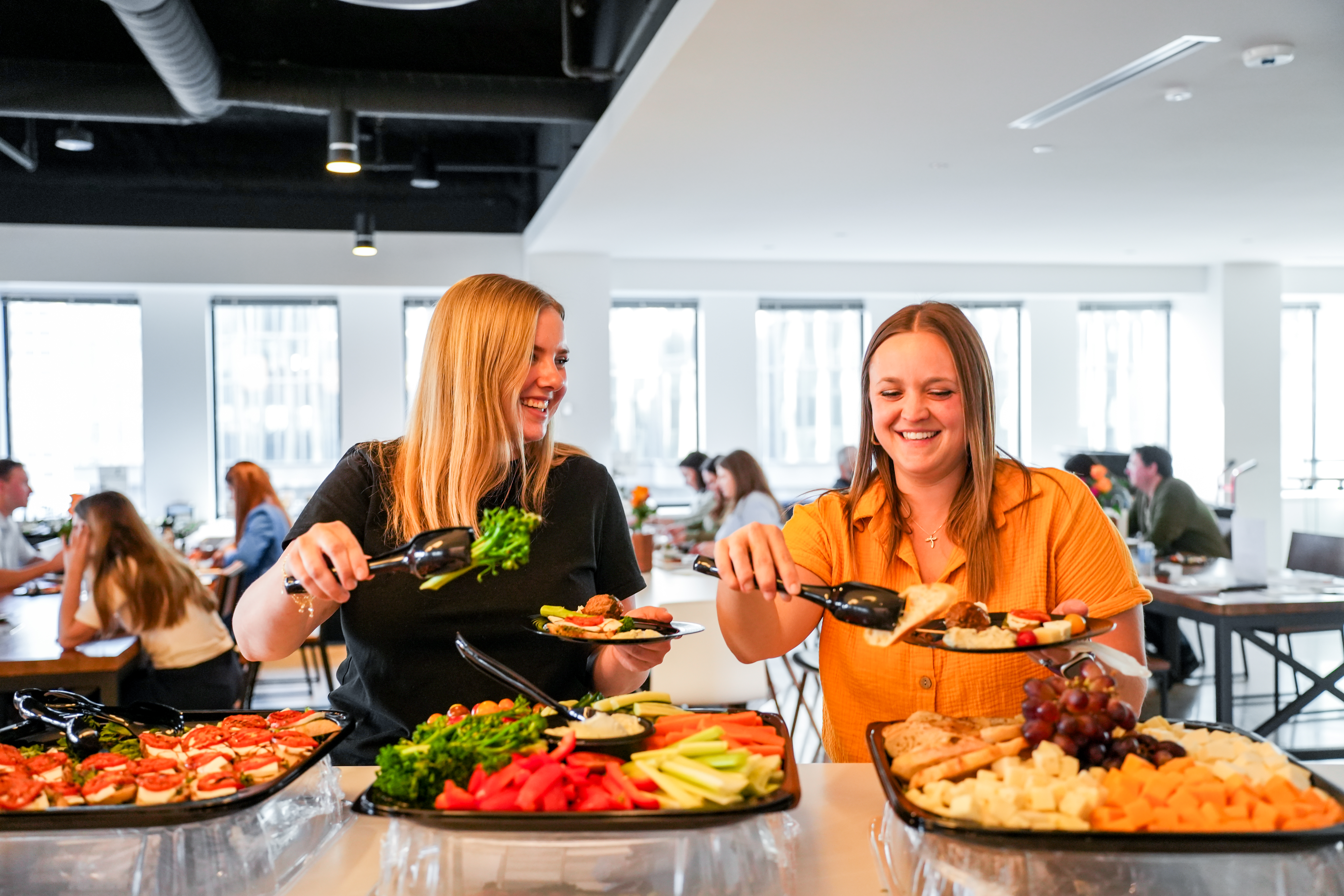 Two smiling coworkers filling their plates from catering platters at an office gathering.