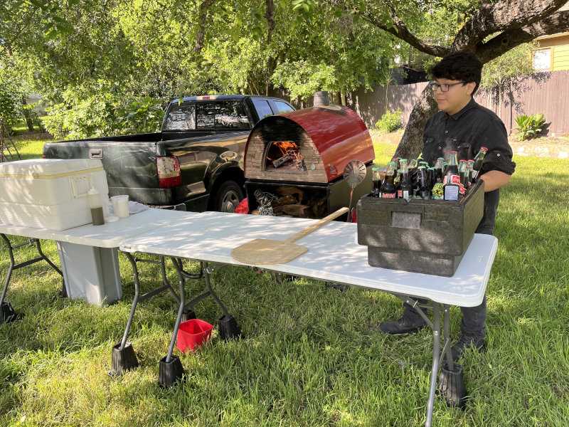 Outdoor cooking setup with a portable pizza oven, cooler, and table with cooking ingredients.