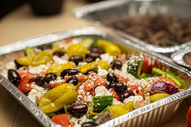 Greek salad in a foil container, featuring olives, cucumbers, tomatoes, feta cheese